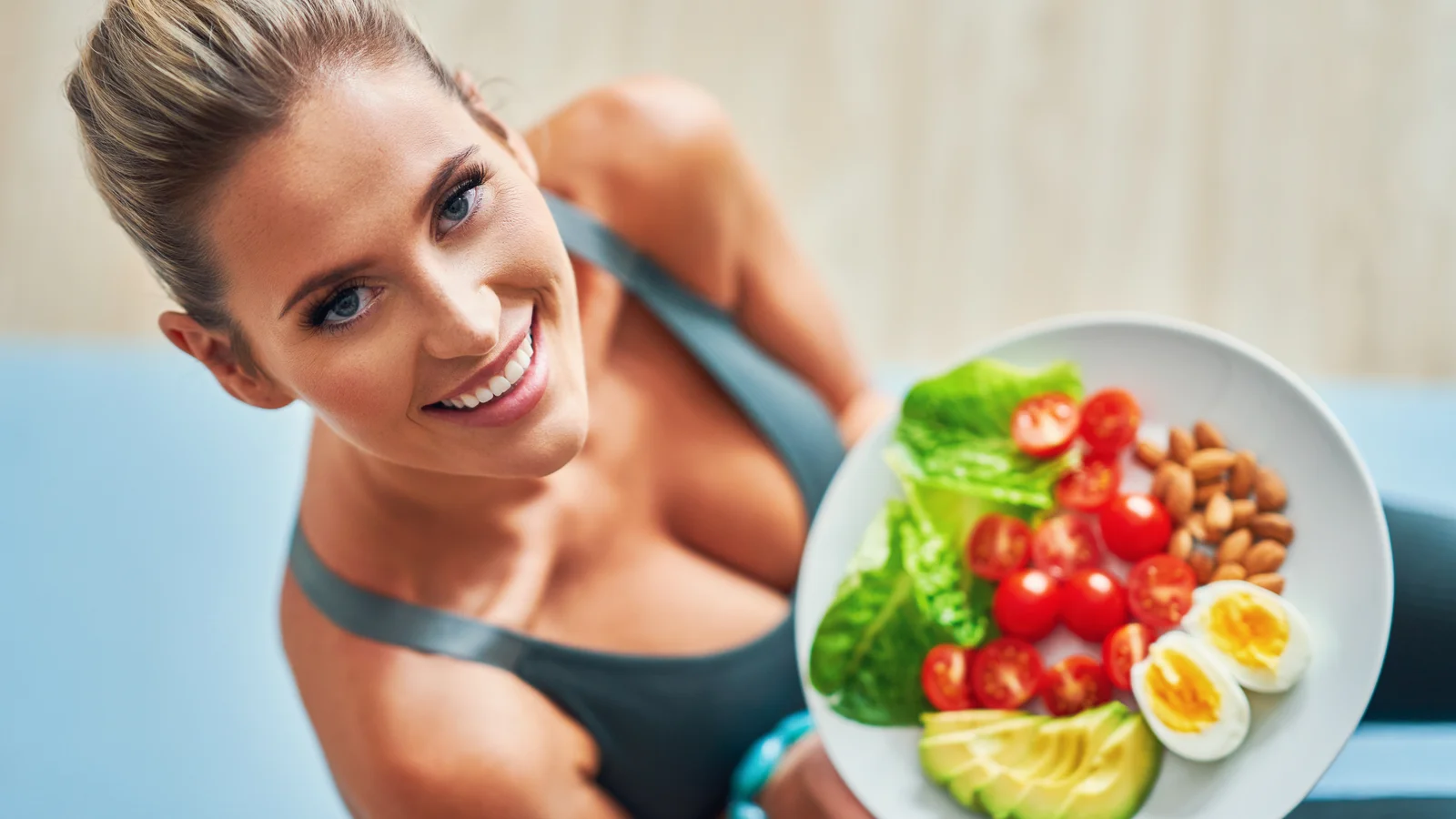 Sportive souriante tenant une assiette équilibrée composée de légumes, œufs, amandes et avocat